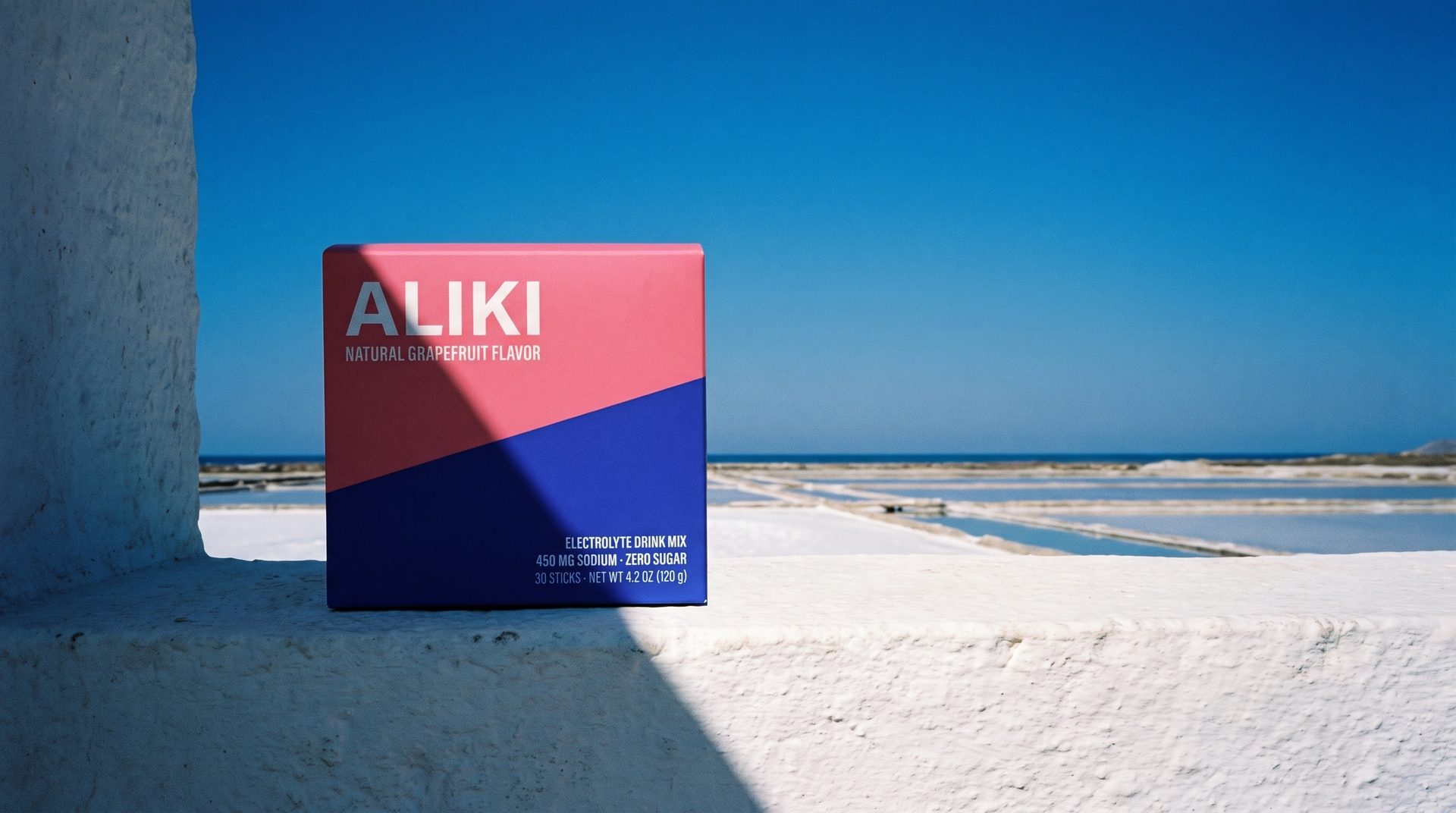 ALIKI carton on a whitewashed ledge in hard sun, with salt pans and the sea behind it