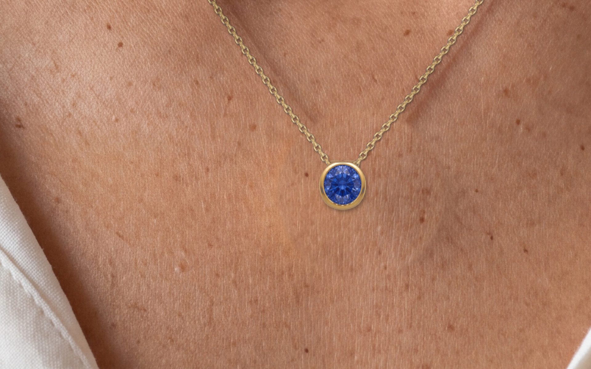 The Corund pendant on a neck: a blue stone in a gold-colored bezel on a fine chain at the collarbones, the edge of a white shirt beside it